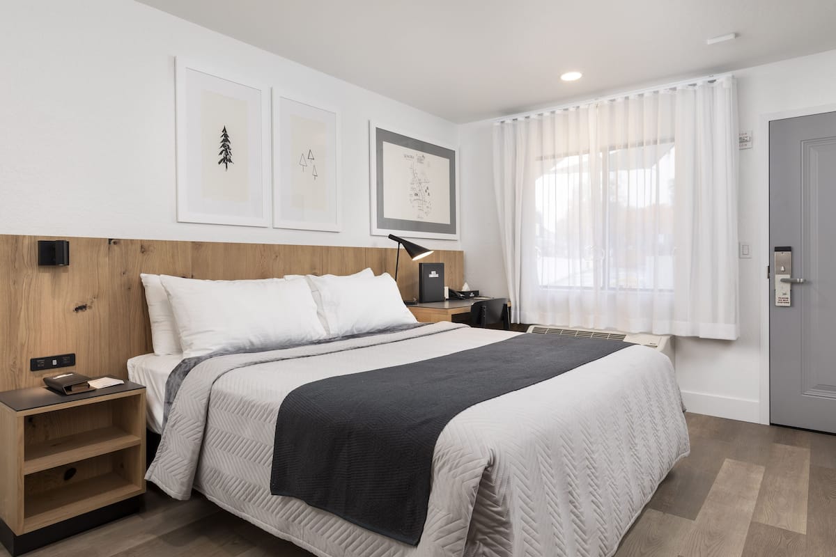 A neatly arranged hotel room with a large bed, a bedside table, a desk, and a window with curtains.