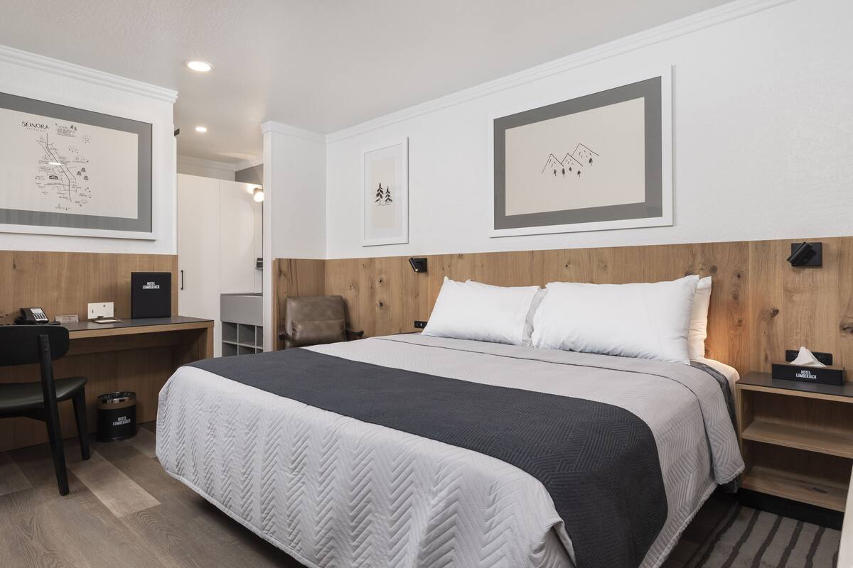 A hotel room with a large bed, a desk, and a chair. There are framed pictures on the wall and a wooden headboard.