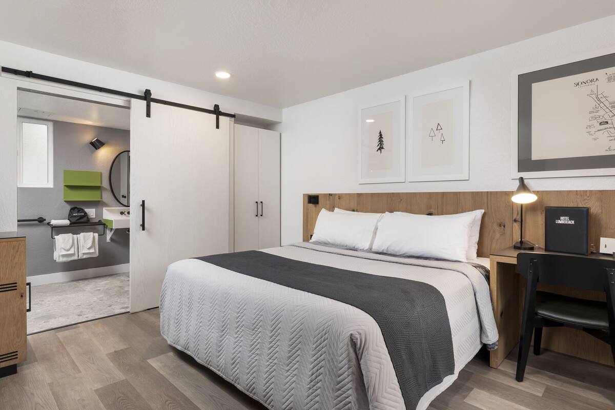 A modern hotel room with a large bed, a wooden headboard, a desk with a chair, and a sliding door leading to a bathroom.