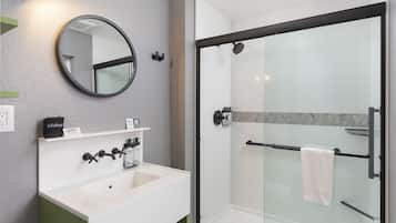 Une salle de bain moderne avec un lavabo blanc, un miroir rond, une douche à parois de verre et un porte-serviettes.