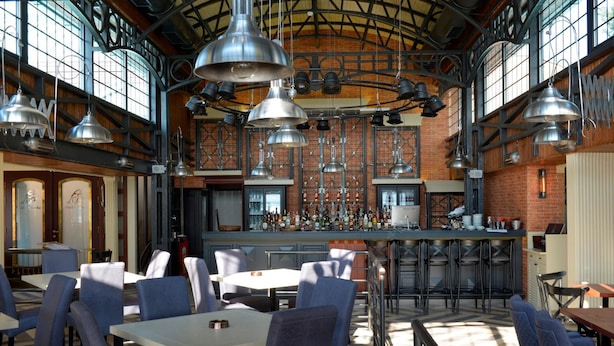 Een ruim restaurant met hoge plafonds, onbehandelde bakstenen muren en diverse zitmogelijkheden, zoals tafels en stoelen.