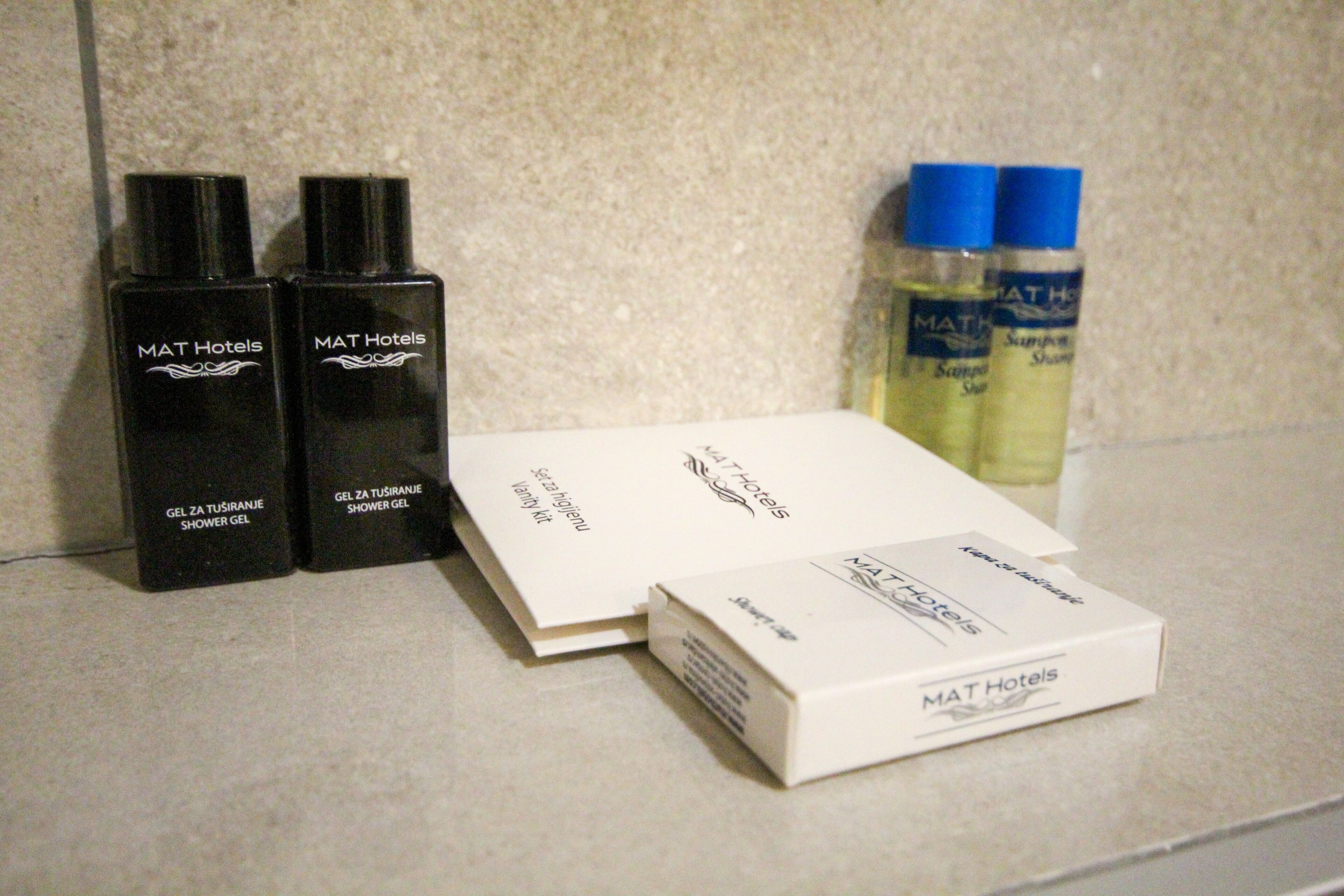 A bathroom counter with toiletries including two black bottles labeled 'MAT Hotels Shower Gel', a white box labeled 'MAT Hotels', and two bottles of shampoo.