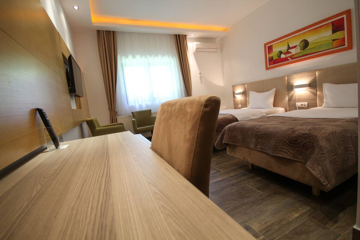 A hotel room with a wooden desk, two beds with headboards, a television mounted on the wall, and a painting above the beds.
