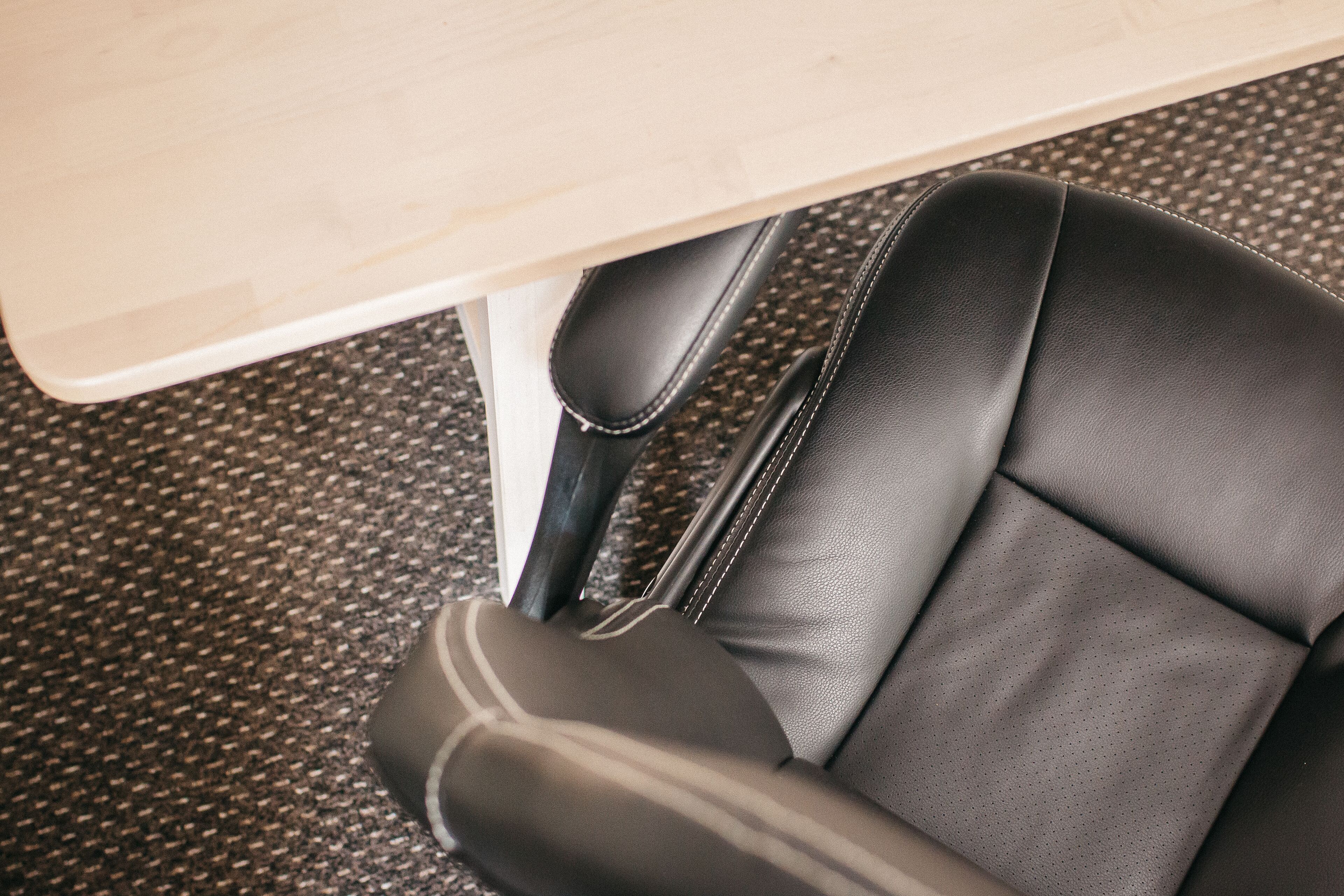 Primer plano de una silla de oficina de cuero negro con costuras visibles y un respaldo perforado, ubicada debajo de una mesa de color claro.