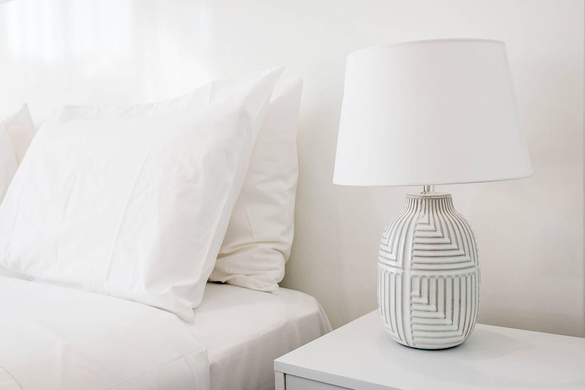 Una cama blanca con un almohadón y una mesita de noche blanca con una lámpara.