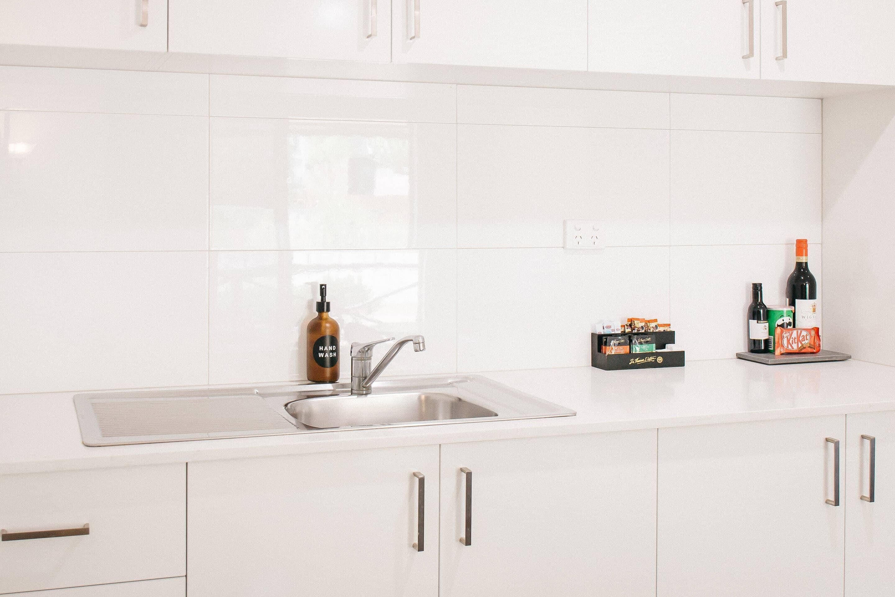 A modern kitchen with white cabinets, a stainless steel sink, and a countertop with various items including a bottle of hand wash soap, a bottle of wine, and a KitKat bar.