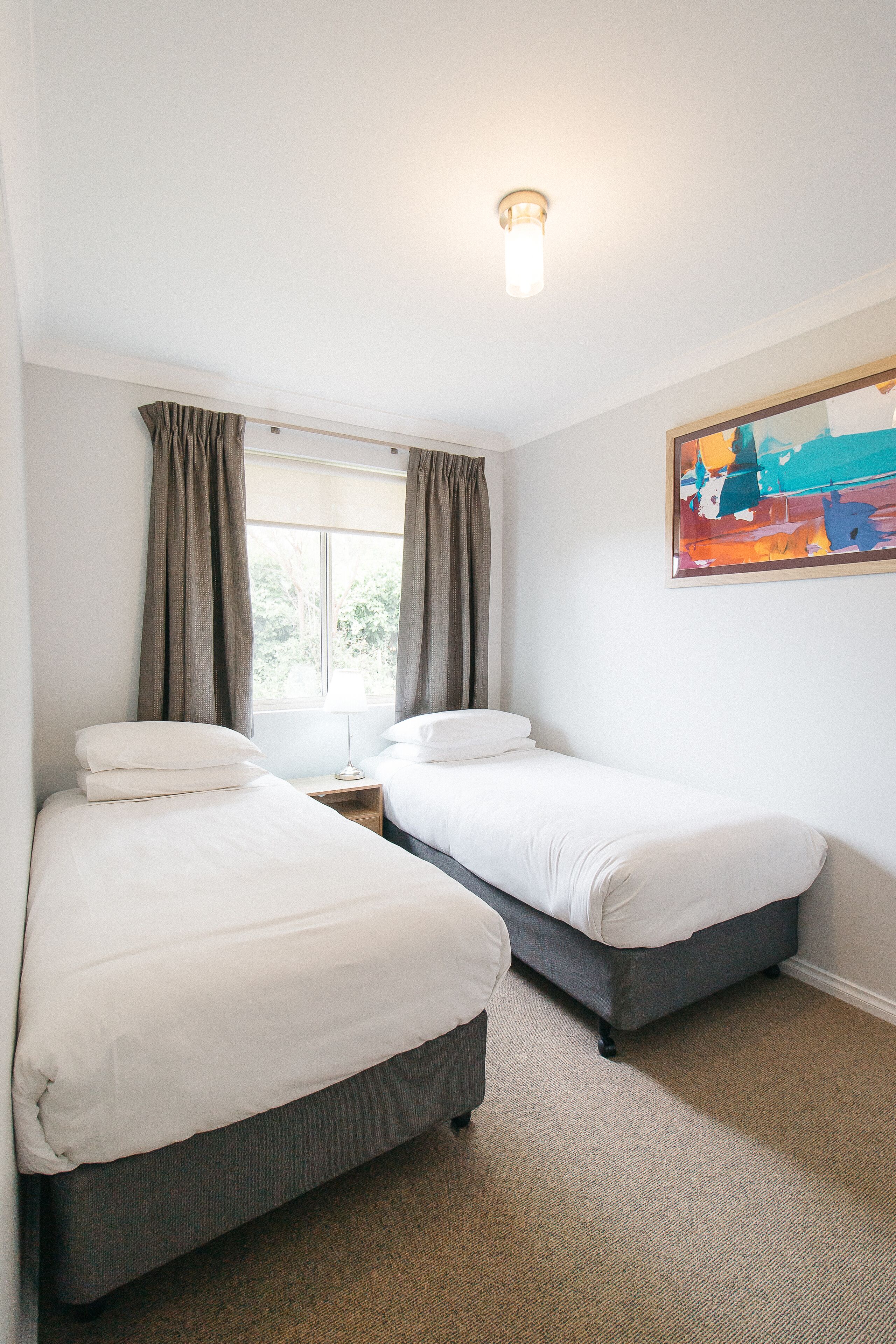 A hotel room with two single beds, a window with curtains, a nightstand, and a painting on the wall.