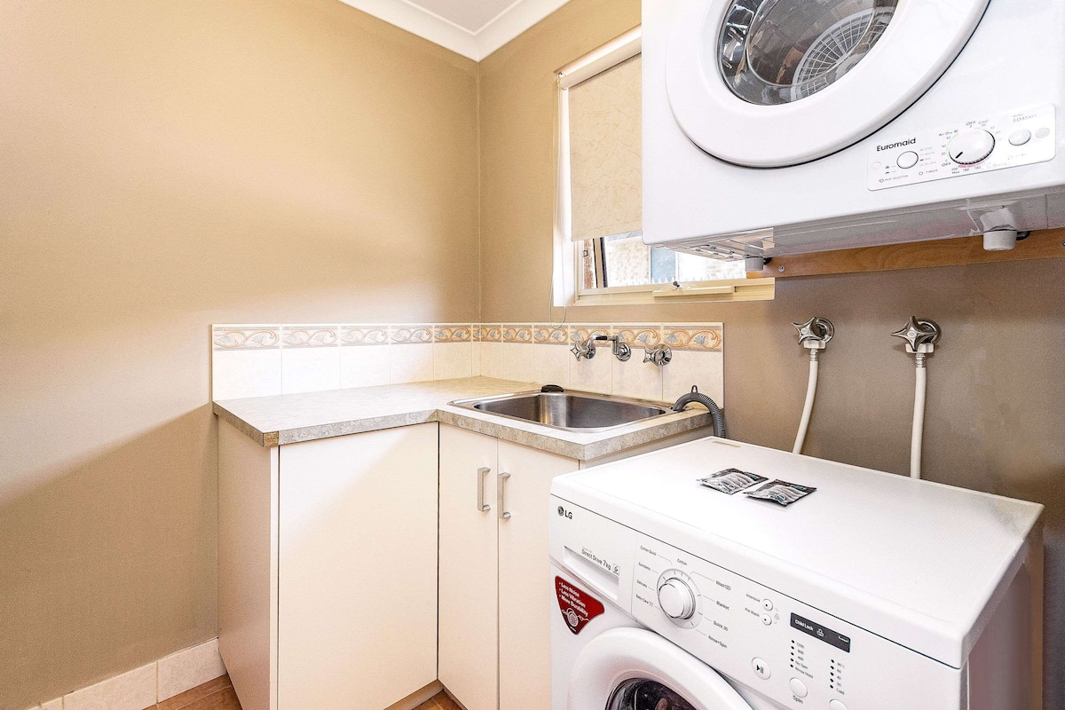 Une petite buanderie équipée d’un lave-linge, d’un évier et d’une fenêtre située au-dessus du lave-linge.