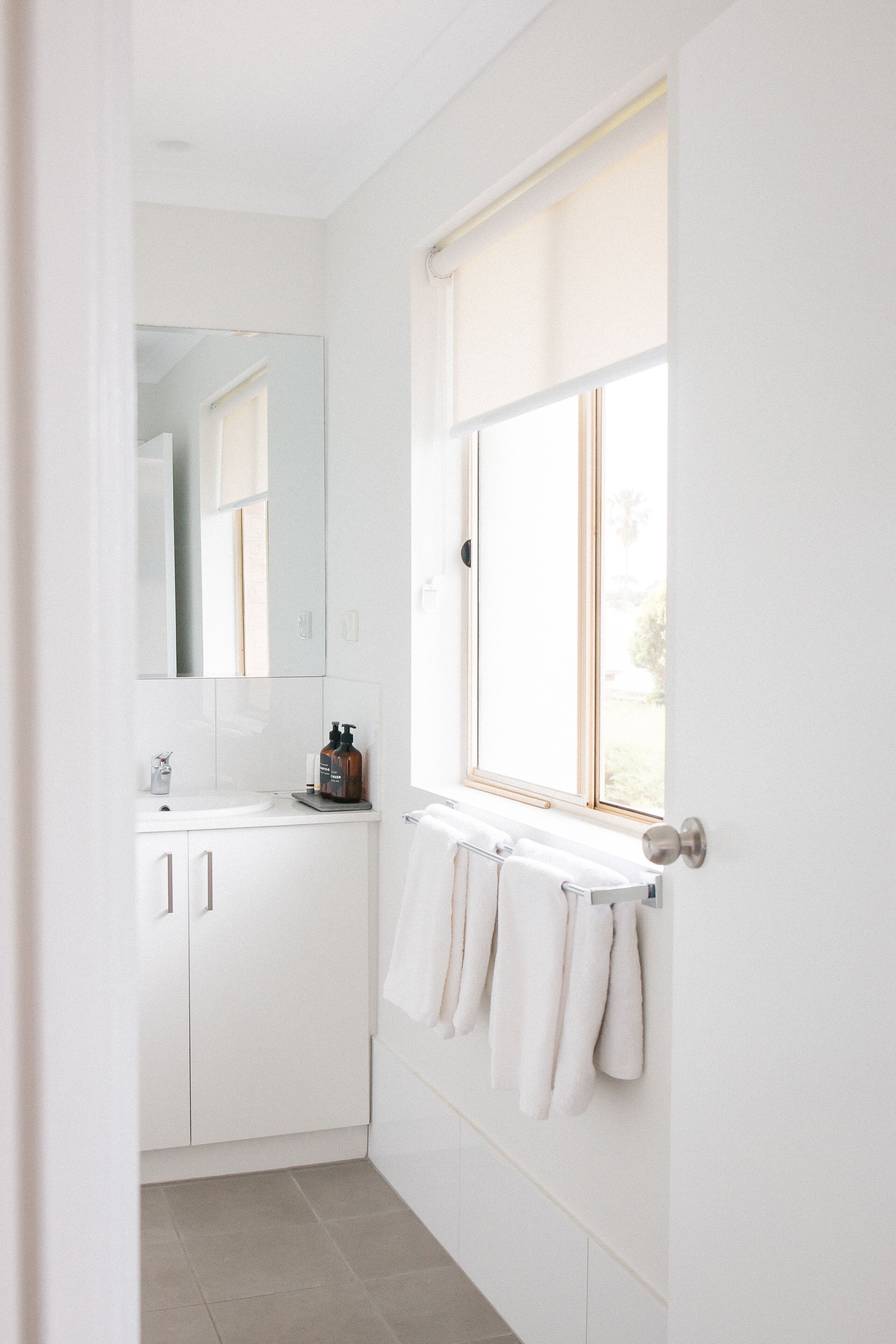 Une salle de bain avec des meubles blancs, un miroir, une fenêtre avec des stores et un porte-serviettes.