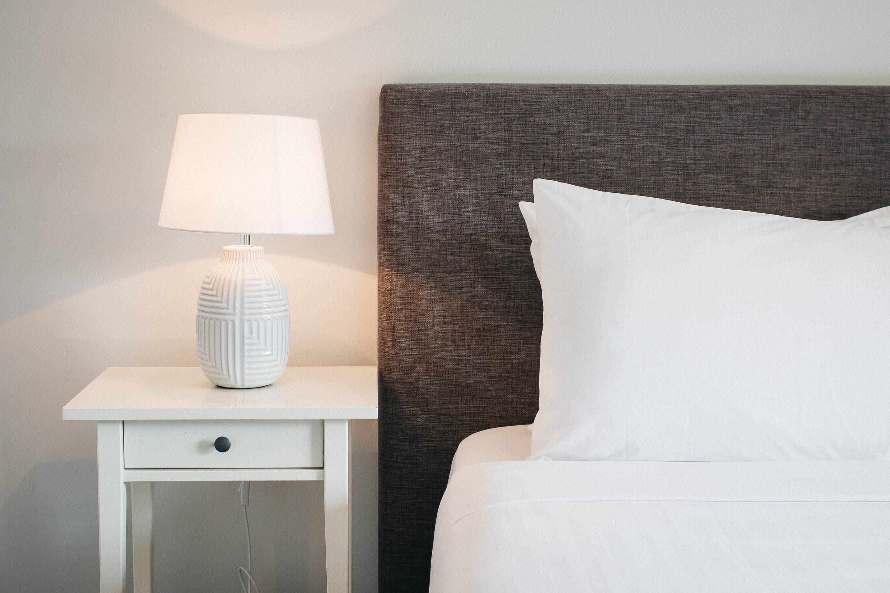 A neatly made bed with white bedding, a white nightstand with a lamp, and a textured headboard.