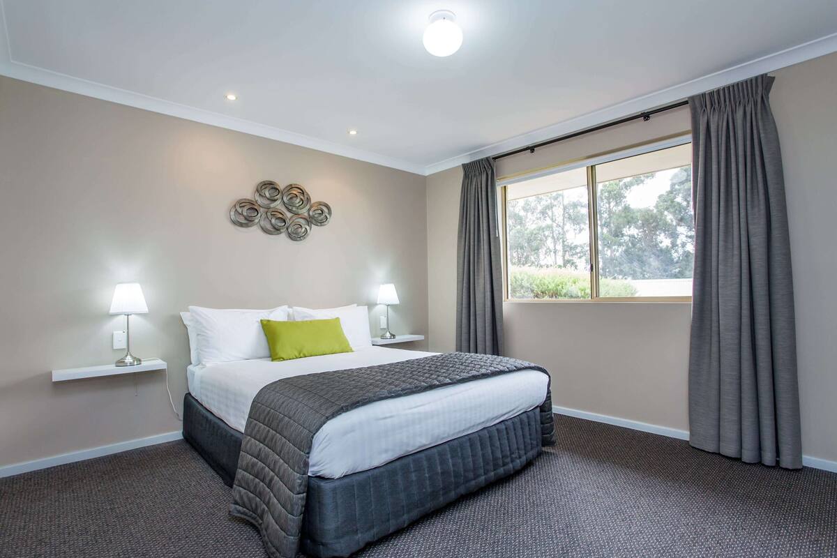 Une chambre d’hôtel avec un lit, deux lampes de chevet, une fenêtre avec des rideaux, un luminaire fixé au mur et une petite étagère avec une lampe.