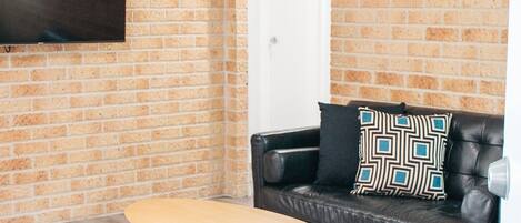 A living room with a brick wall, a flat-screen TV mounted above a wooden coffee table, a black leather sofa with patterned cushions, and a white door in the background.