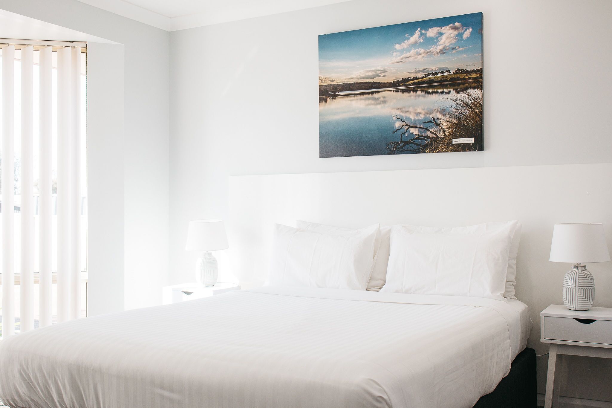 A hotel room with a large bed, white bedding, a nightstand with a lamp, and a wall-mounted picture of a lake.