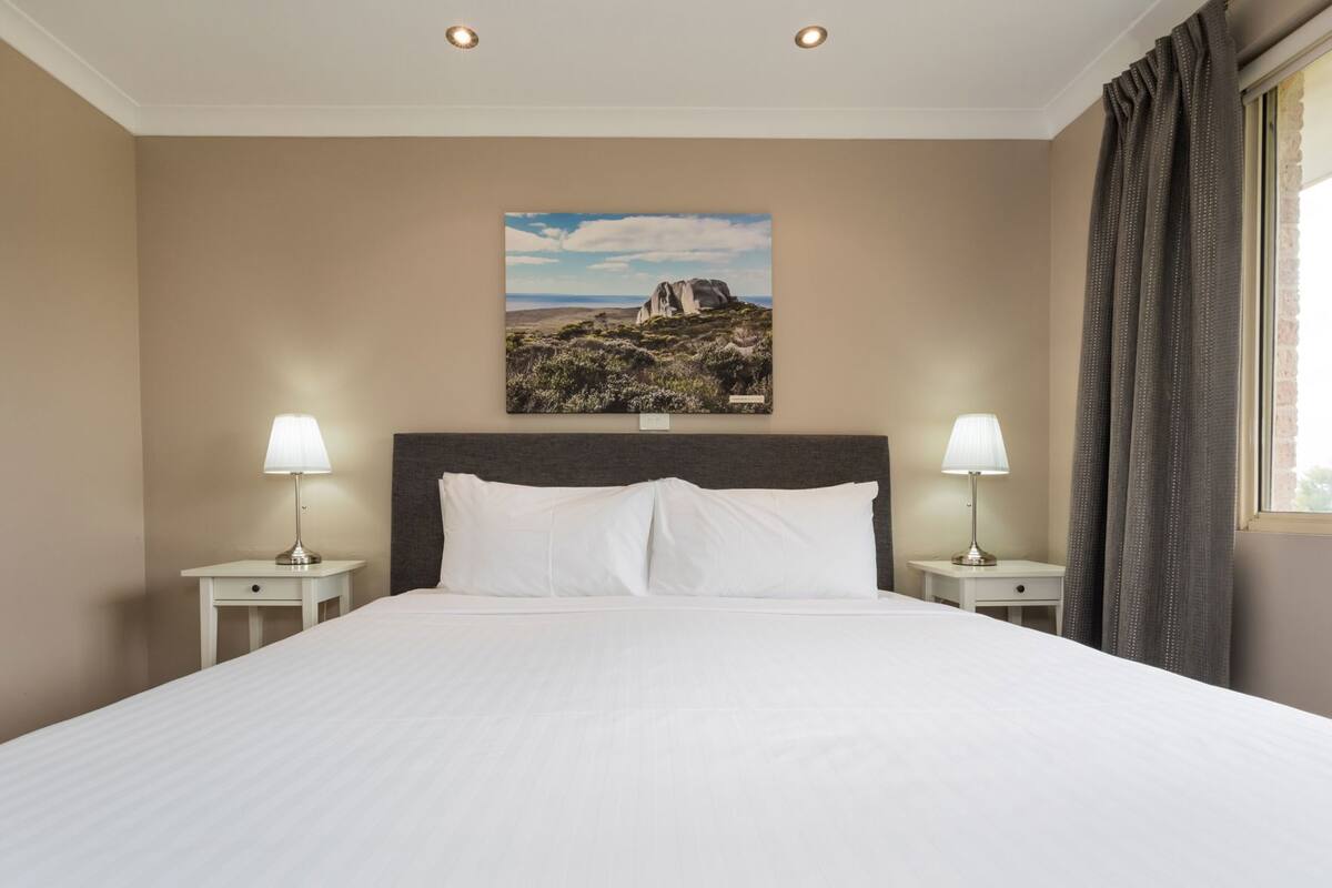 Une chambre d’hôtel avec un grand lit, deux tables de chevet avec des lampes, un tableau au mur et une fenêtre avec des rideaux.