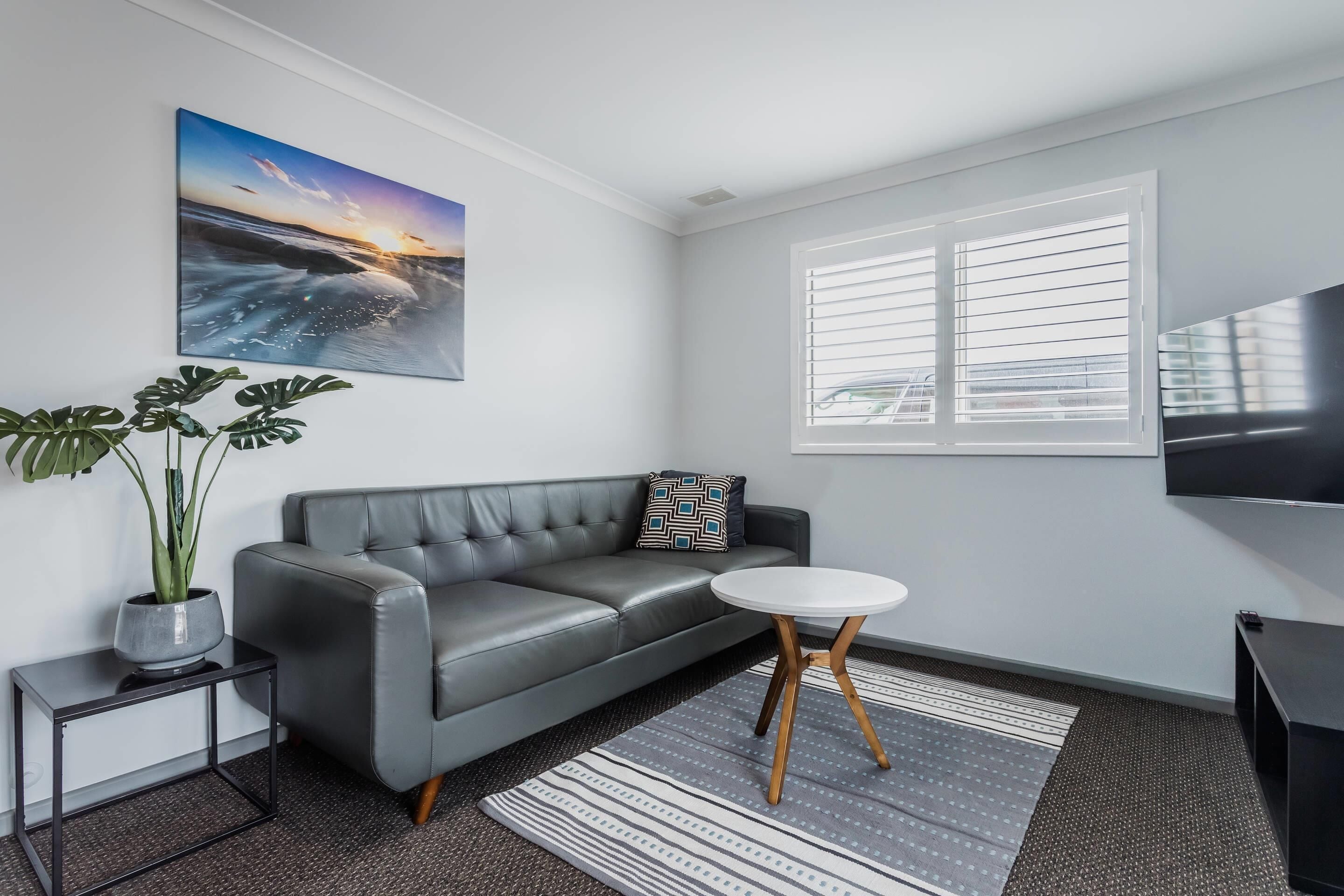 A modern living room with a grey leather sofa, a small white table, a potted plant, a striped rug, a large window with blinds, and a flat-screen TV mounted on the wall.