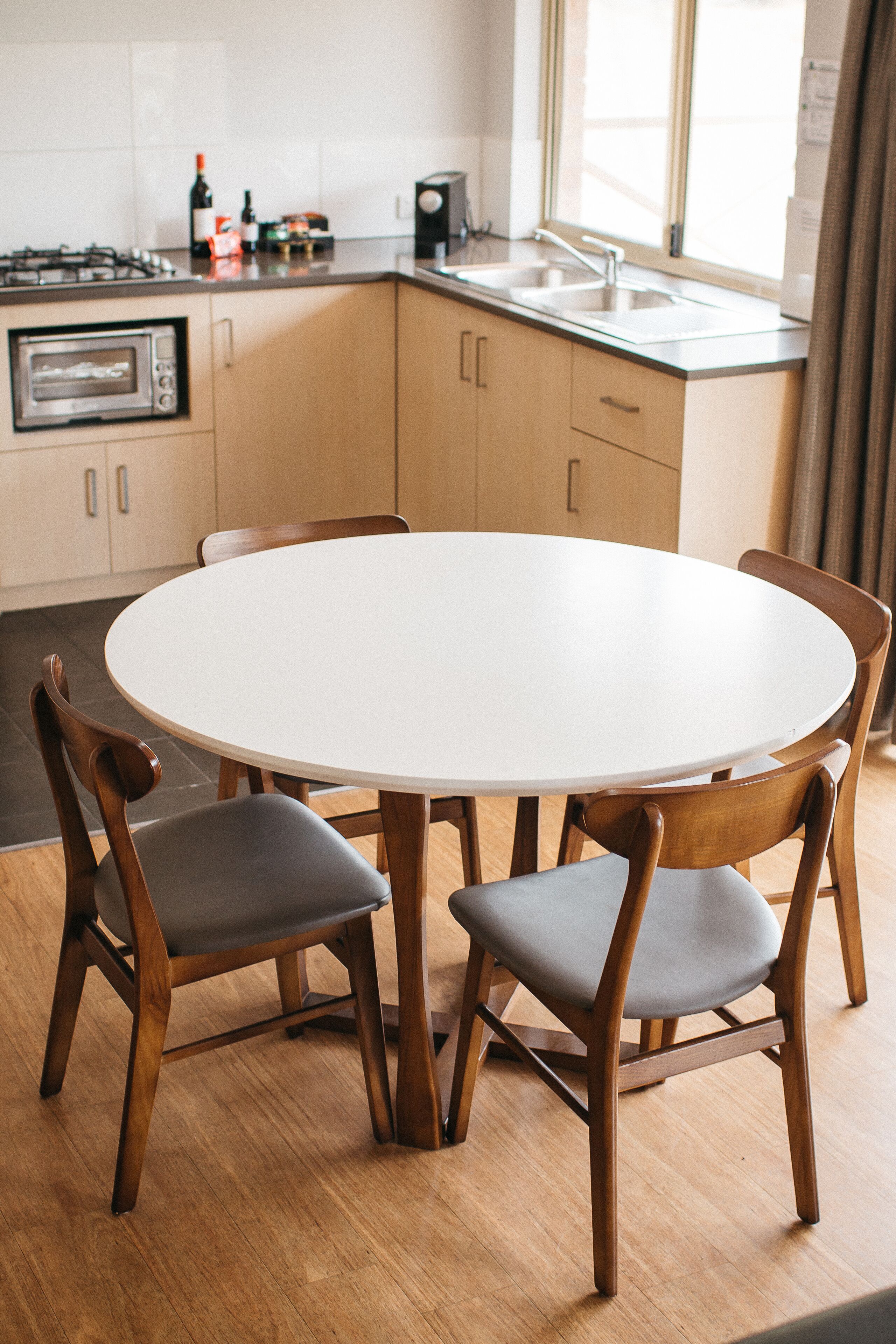 A modern kitchen with a round dining table, wooden chairs, a built-in oven, and a window with curtains.