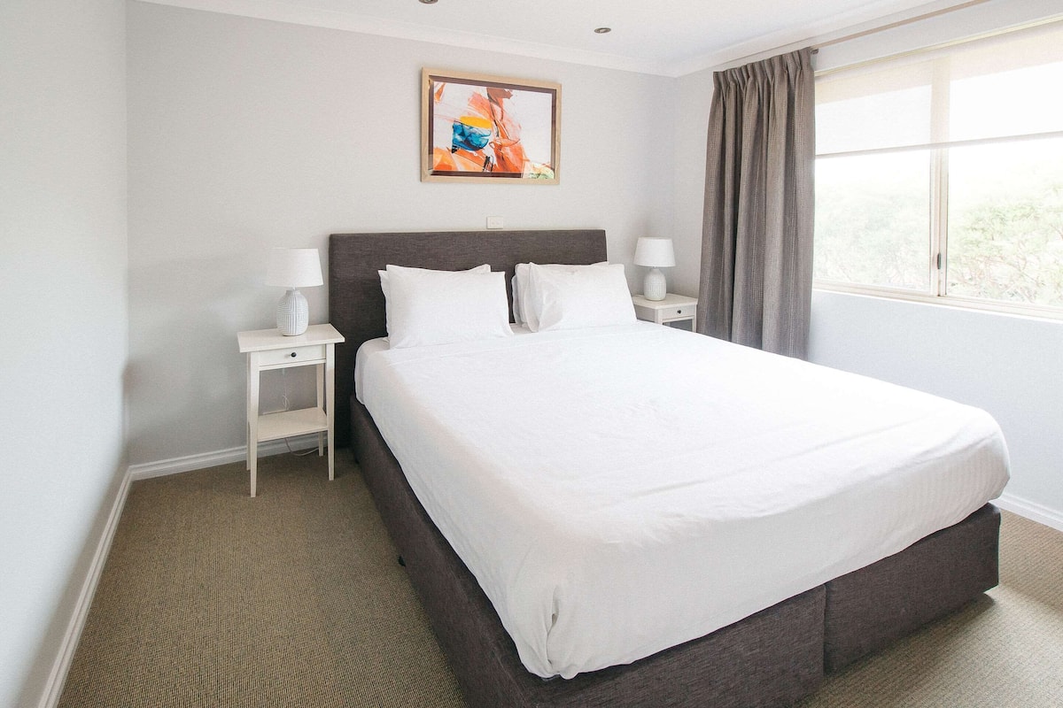 Un lit bien fait dans une chambre d’hôtel, avec une grande fenêtre, un tableau au mur et une table de chevet avec une lampe.