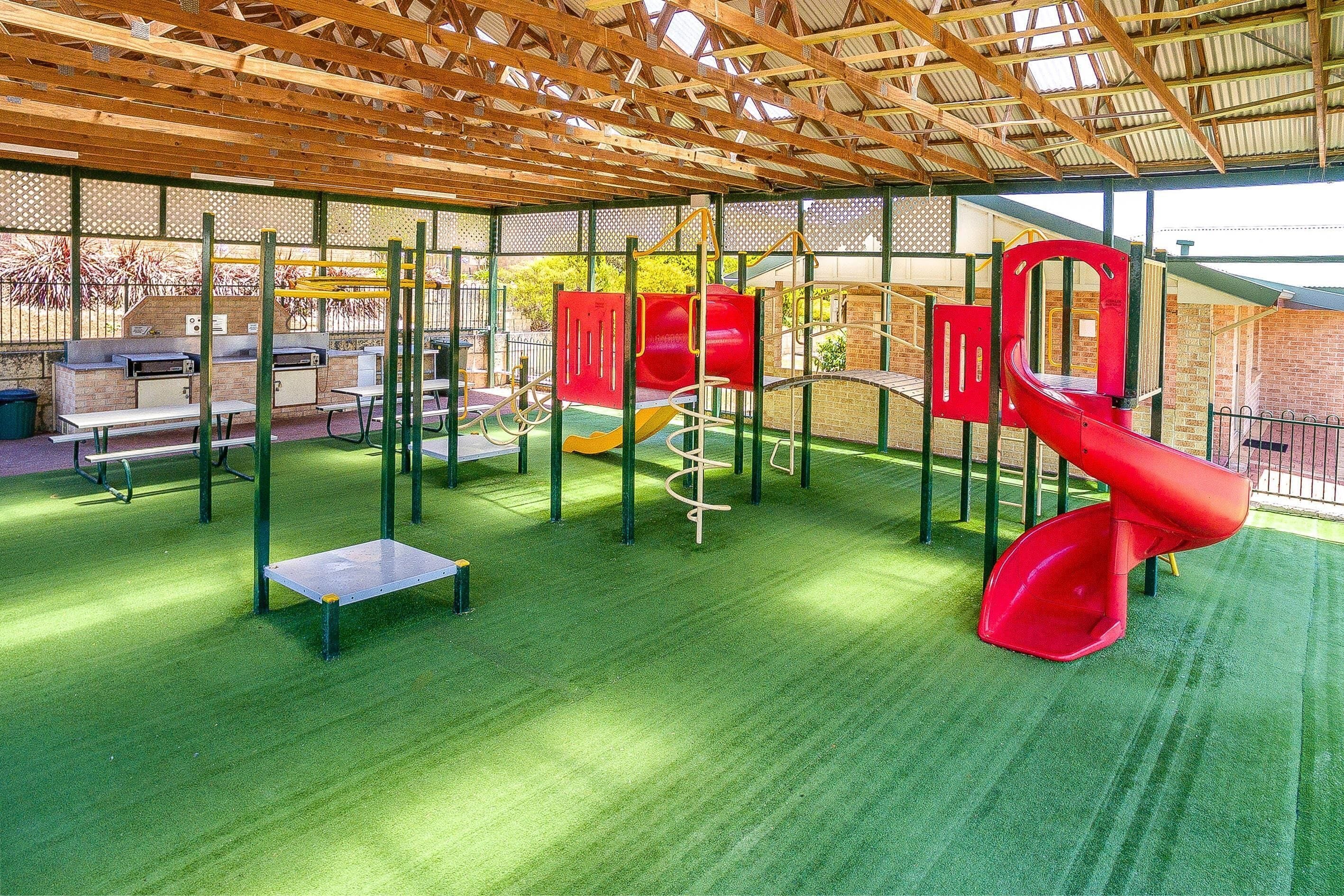 A playground with a red slide, climbing structures, and a covered area with artificial turf.