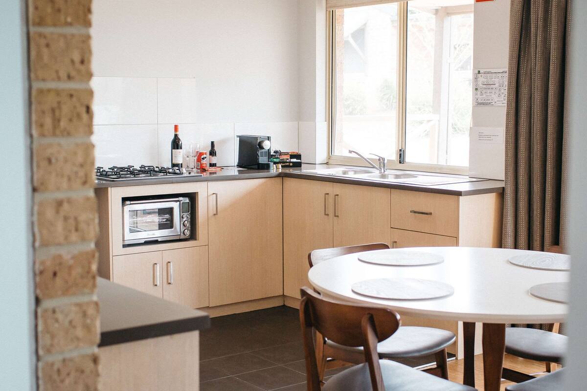 Une cuisine compacte équipée d’un four à micro-ondes, d’une cuisinière à gaz et d’une machine à café. On y trouve une table à manger avec des chaises, ainsi qu’une fenêtre avec des rideaux.