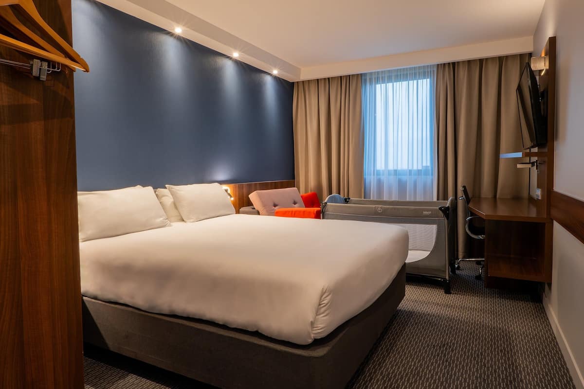 A hotel room with a large bed, a desk, a chair, a TV, and a window with curtains.