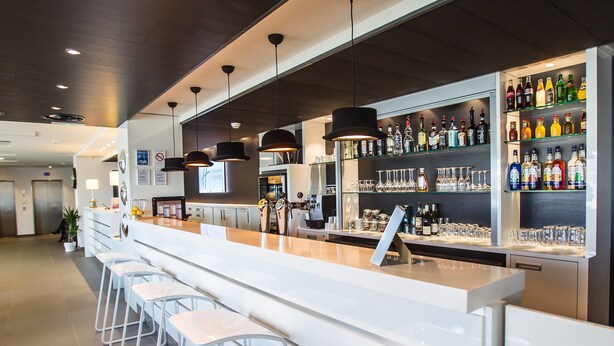 A modern bar area with a long counter, stools, and shelves stocked with bottles and glasses. - Holiday Inn Express Dijon by IHG (Saint-Apollinaire)