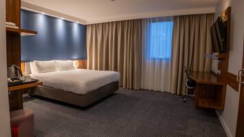 A hotel room with a bed, a desk with a kettle, a chair, a TV, and a window with curtains.