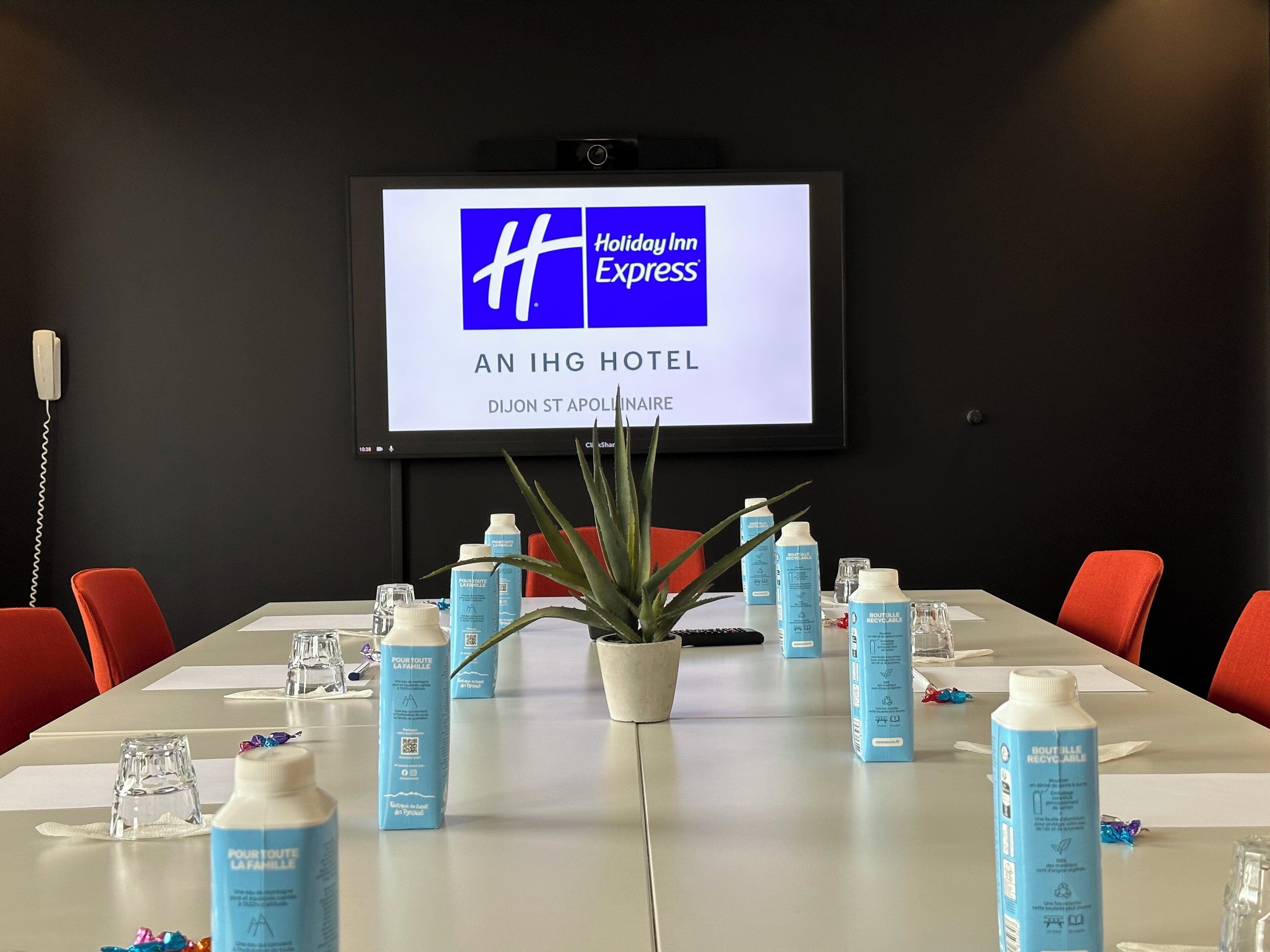 A conference room with a large screen displaying the Holiday Inn Express logo, a table with water bottles and a potted plant, and red chairs around the table.