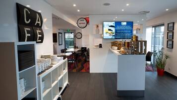 Un moderno vestíbulo de hotel con una zona de café, un mostrador de recepción y una pantalla digital que muestra información sobre el desayuno.