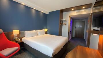 Een hotelkamer met een bed, een rode stoel, een bureau met een lamp, een televisie en een deur die naar een andere kamer leidt.