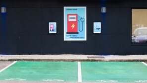 Una estación de carga con un letrero que indica que es gratuita para clientes, un espacio de estacionamiento con un carril bici verde, y un vehículo al fondo.