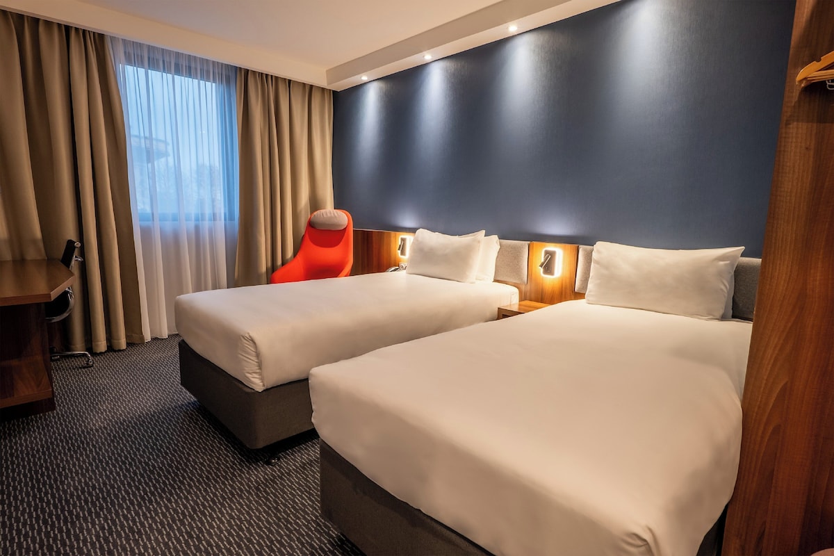 Habitación de hotel con dos camas, un escritorio de madera, una silla y un ventanal con cortinas.