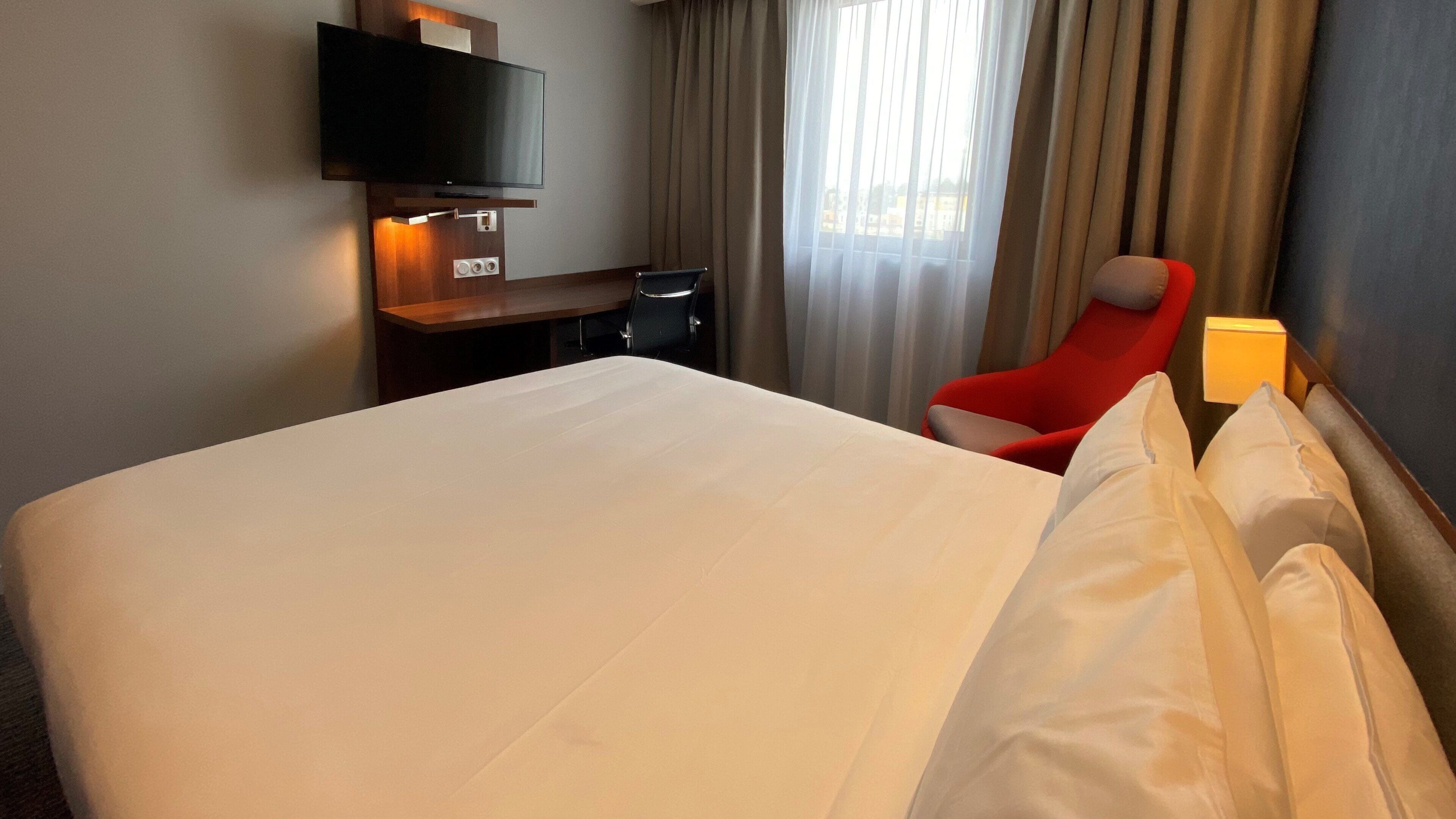 Una habitación de hotel con una cama grande, un escritorio, un televisor y una silla roja.