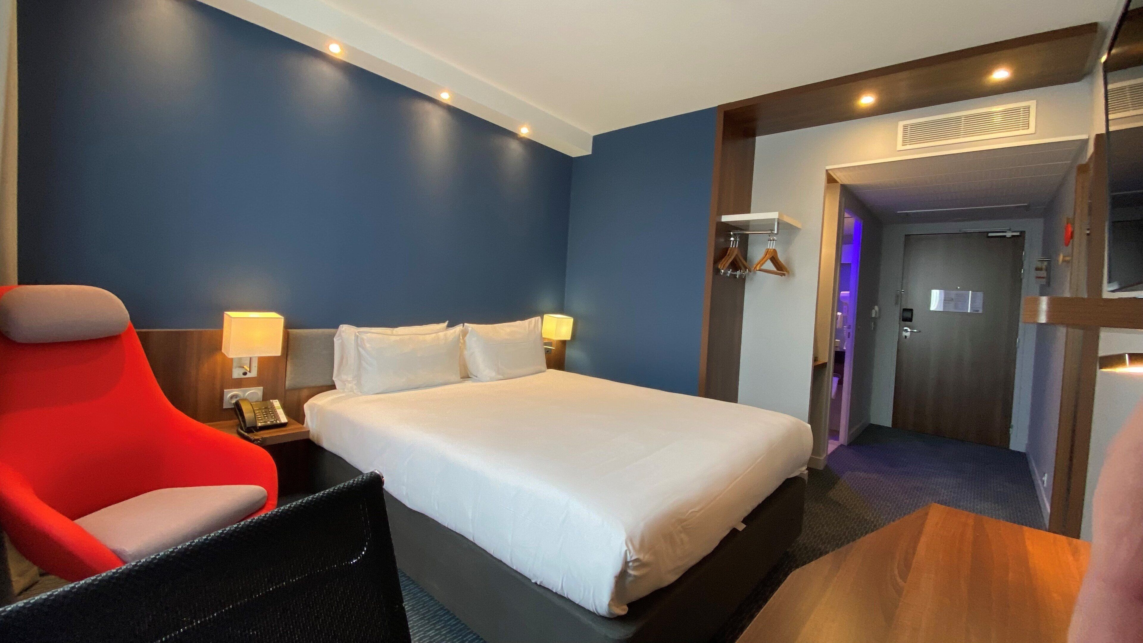 Camera d'albergo con un letto grande, una poltrona rossa, una scrivania con il telefono e una TV a parete.