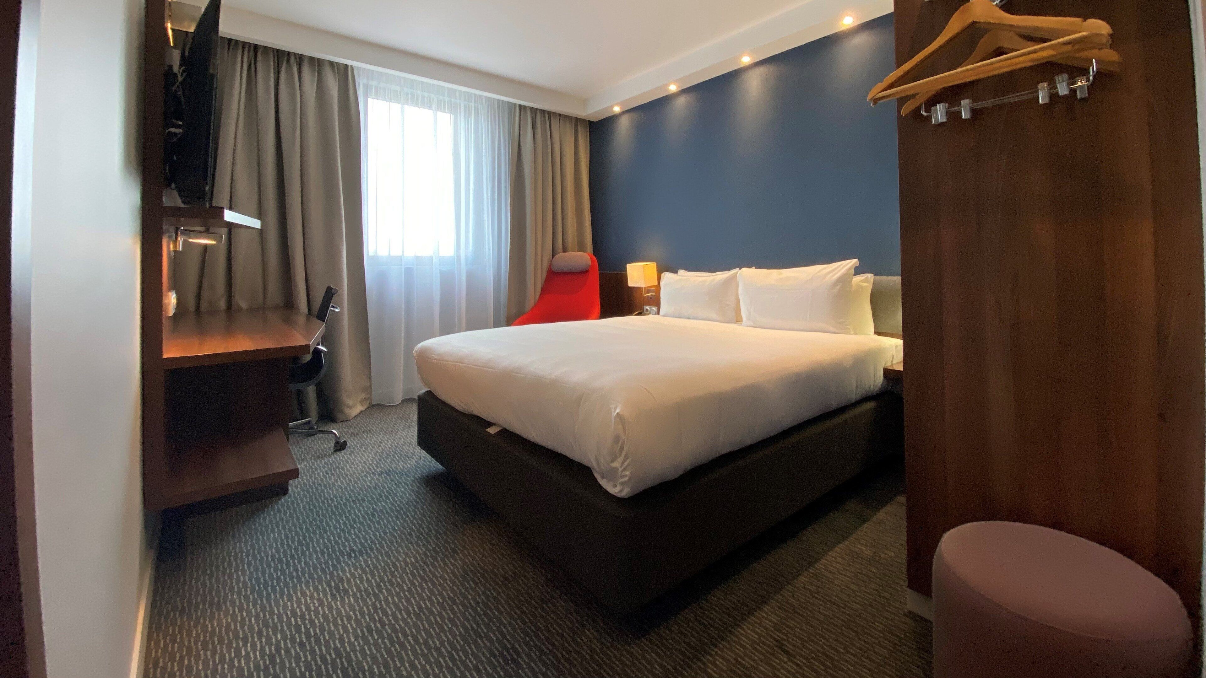 A hotel room with a large bed, a desk, a chair, a TV, and a wooden hanger rack.