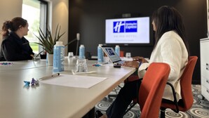 Une salle de conférence équipée d’un grand écran diffusant une présentation, d’une table avec des bouteilles d’eau, d’un ordinateur portable et d’une plante.