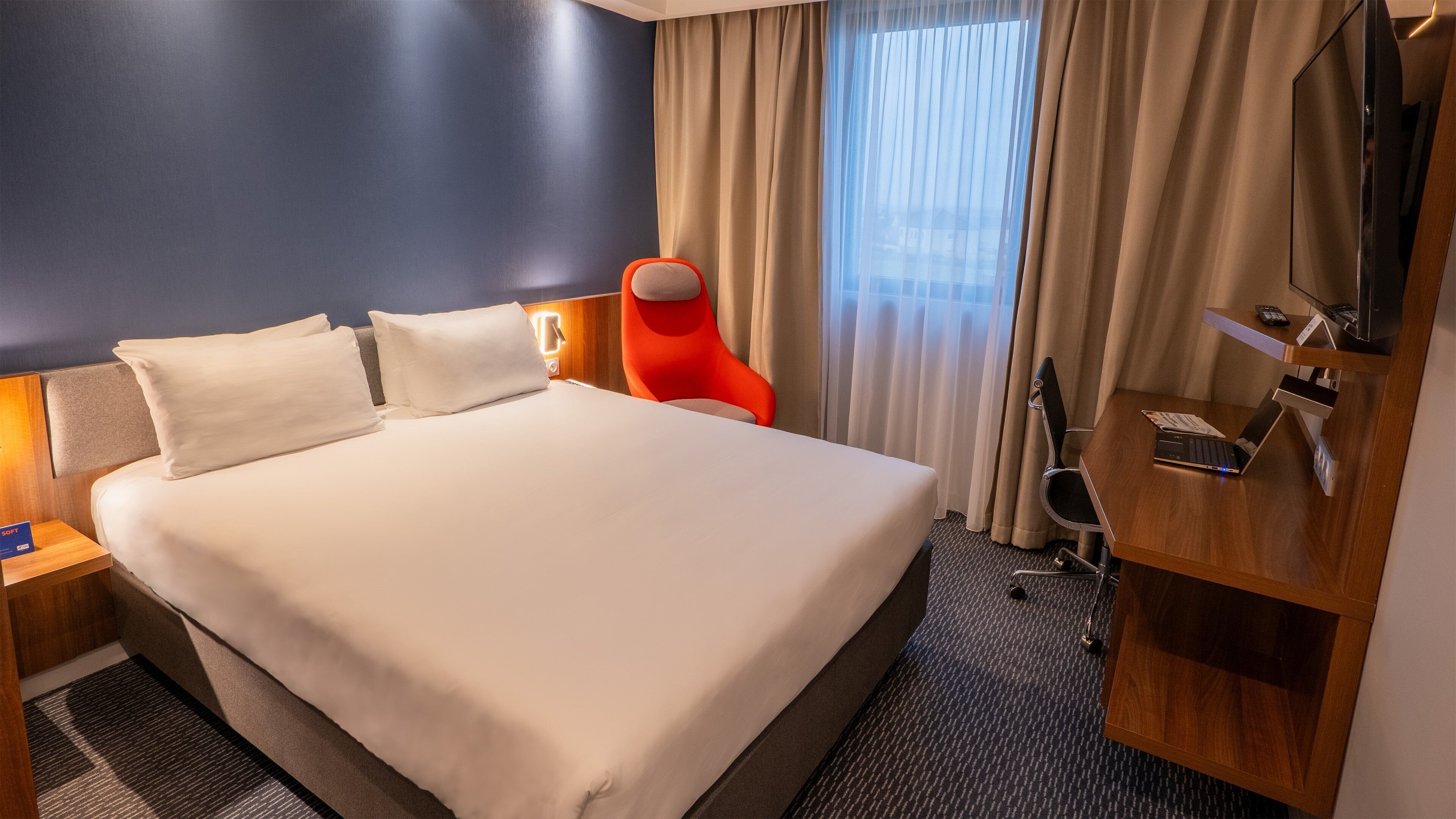A hotel room with a large bed, a red chair, a desk with a laptop, and a mirror.