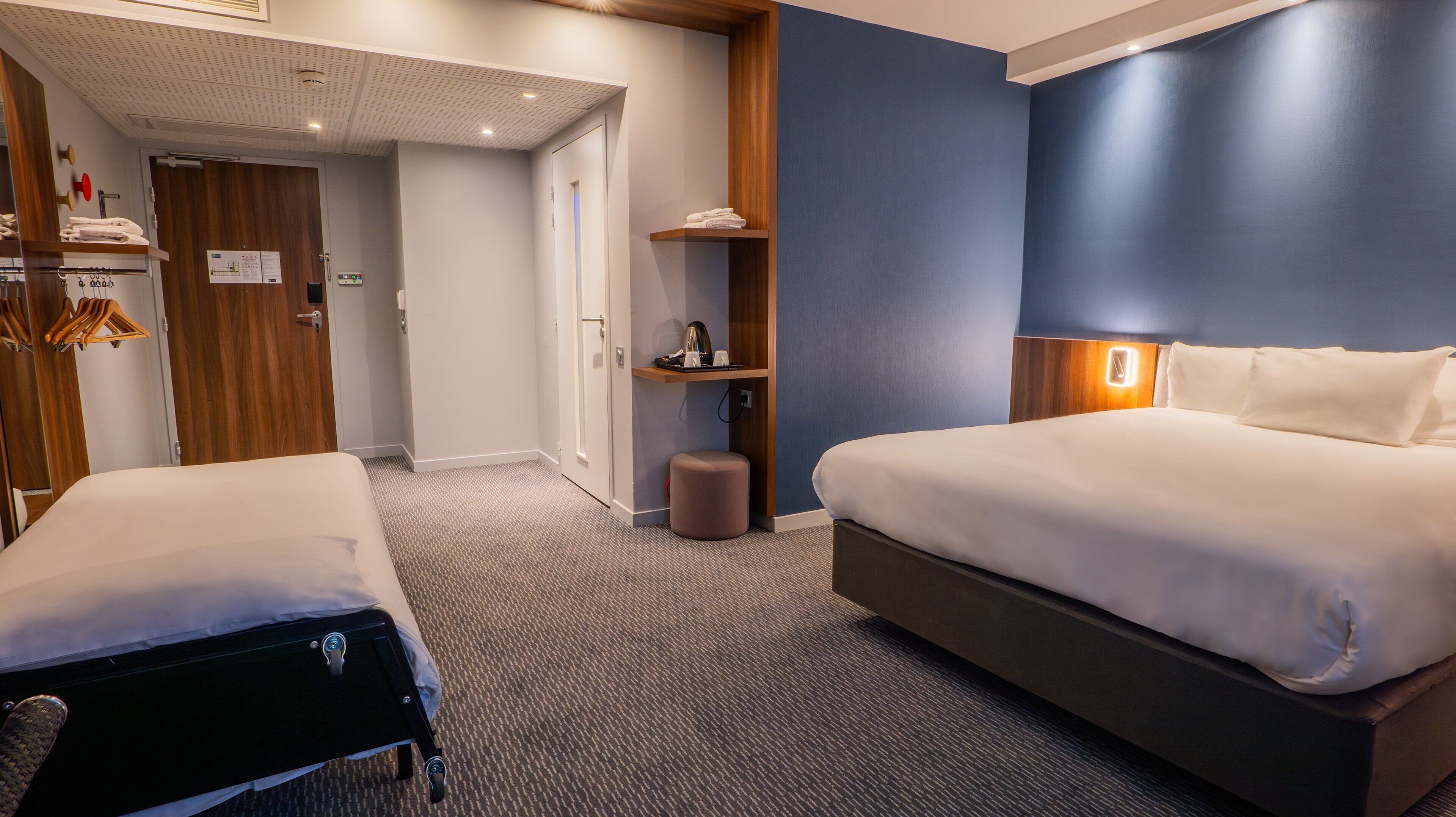 A hotel room with a large bed, a smaller bed, a wardrobe, a mirror, a shelf with towels, and a small stool.