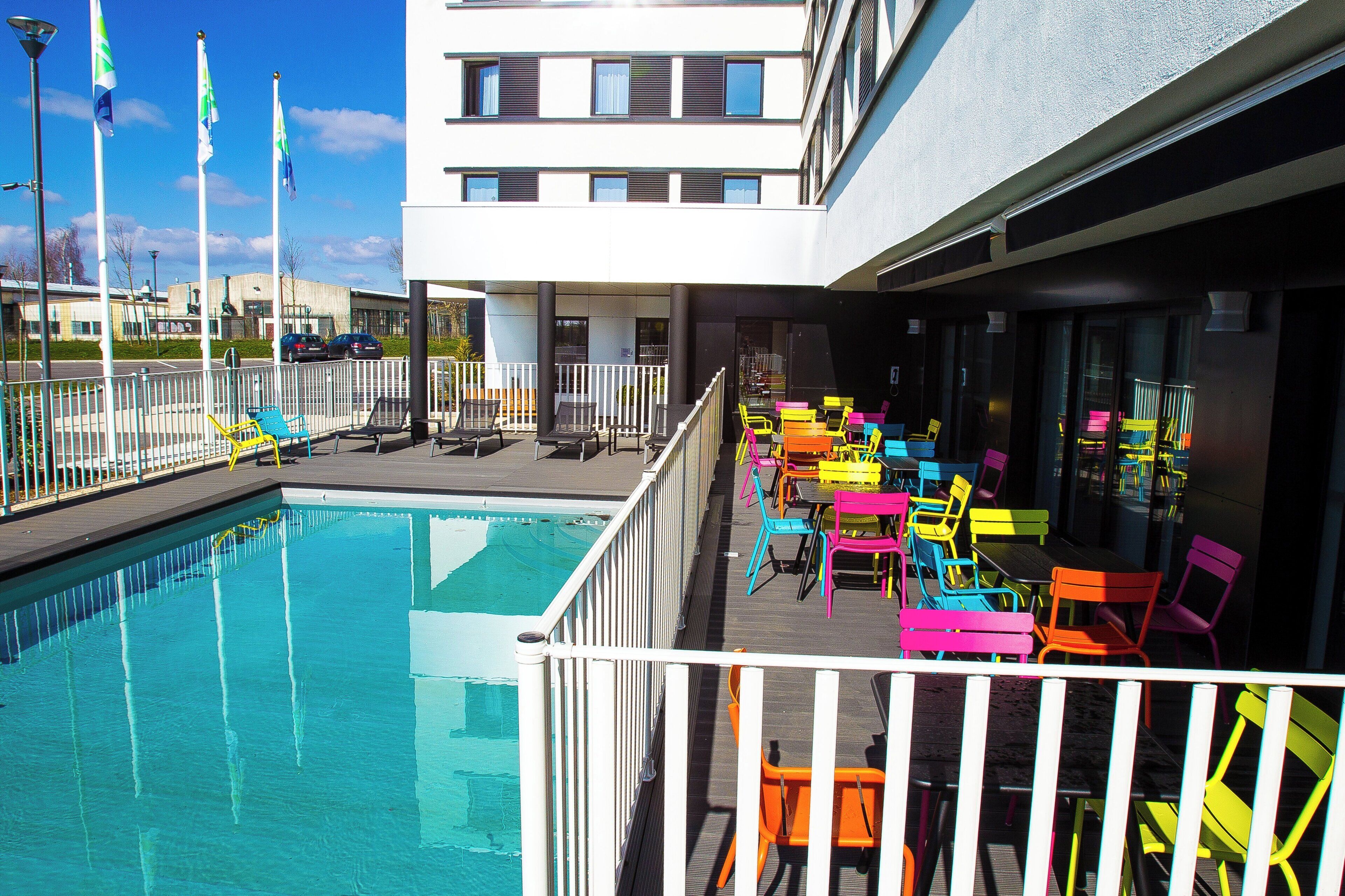 Piscina com área de deck, mobiliário de exterior com cadeiras coloridas e um edifício com várias janelas ao fundo.