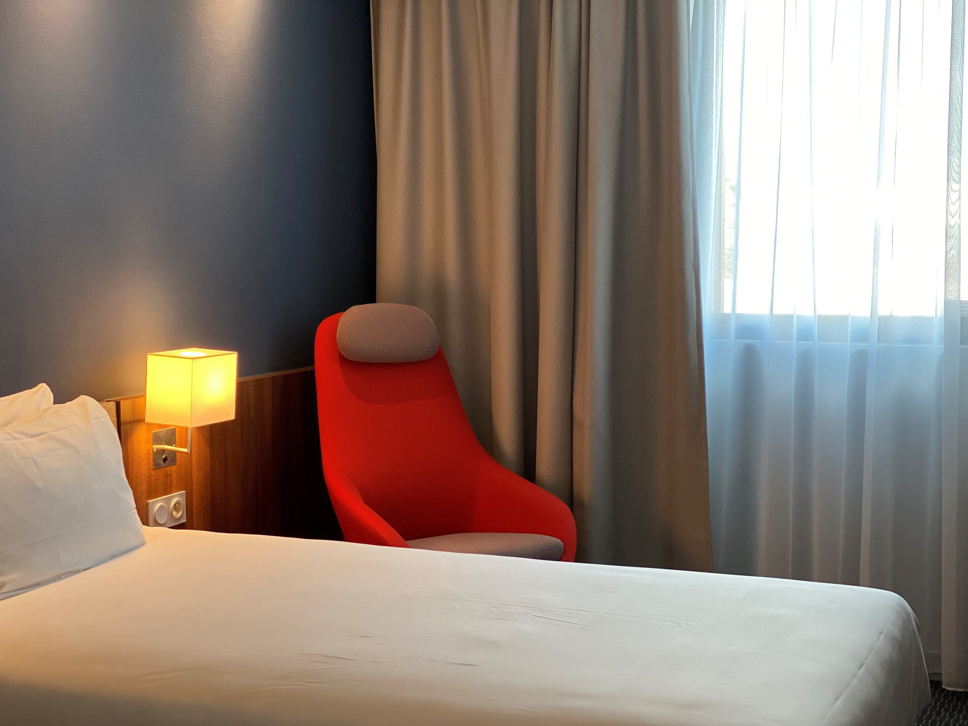 A hotel room with a bed, a red armchair, a lamp, and a window with curtains.