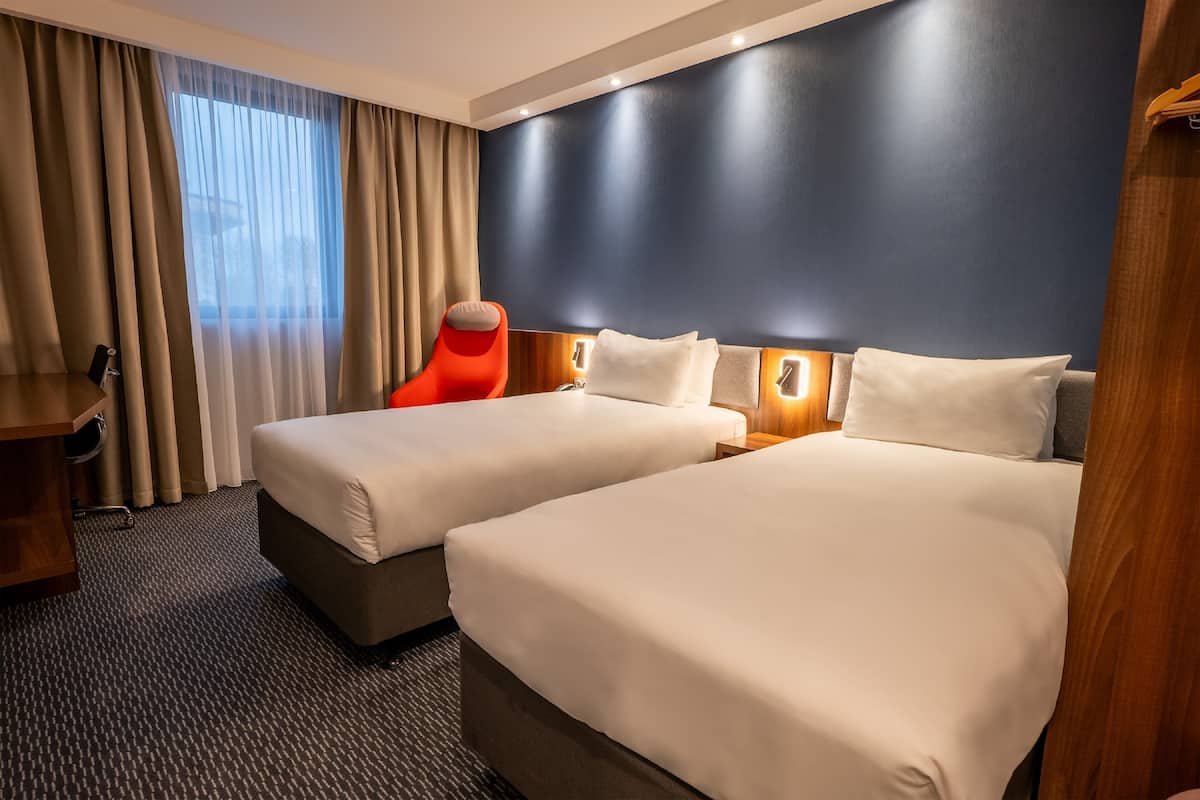 A hotel room with two beds, a wooden desk, a chair, and a large window with curtains.