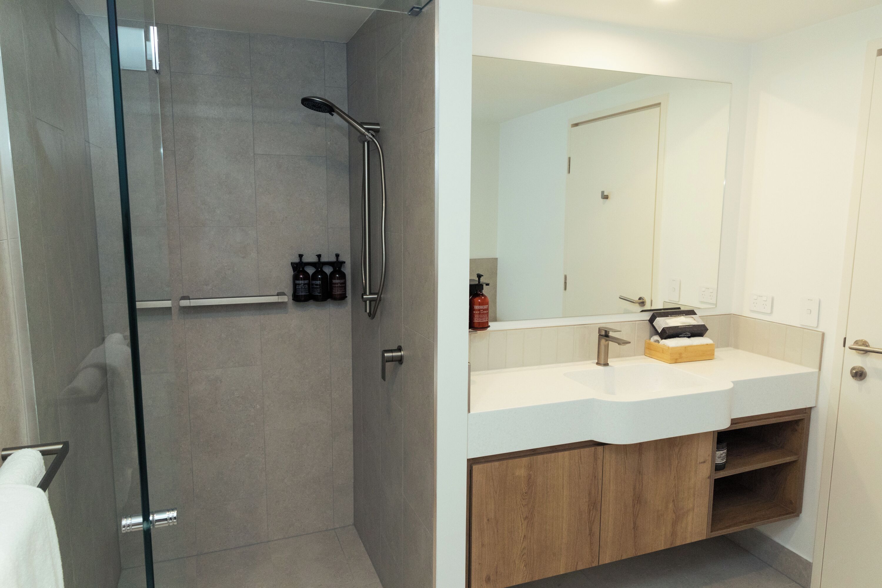 Un baño moderno con ducha envuelta en vidrio, lavamanos blanco con mueble de madera y un espejo grande.