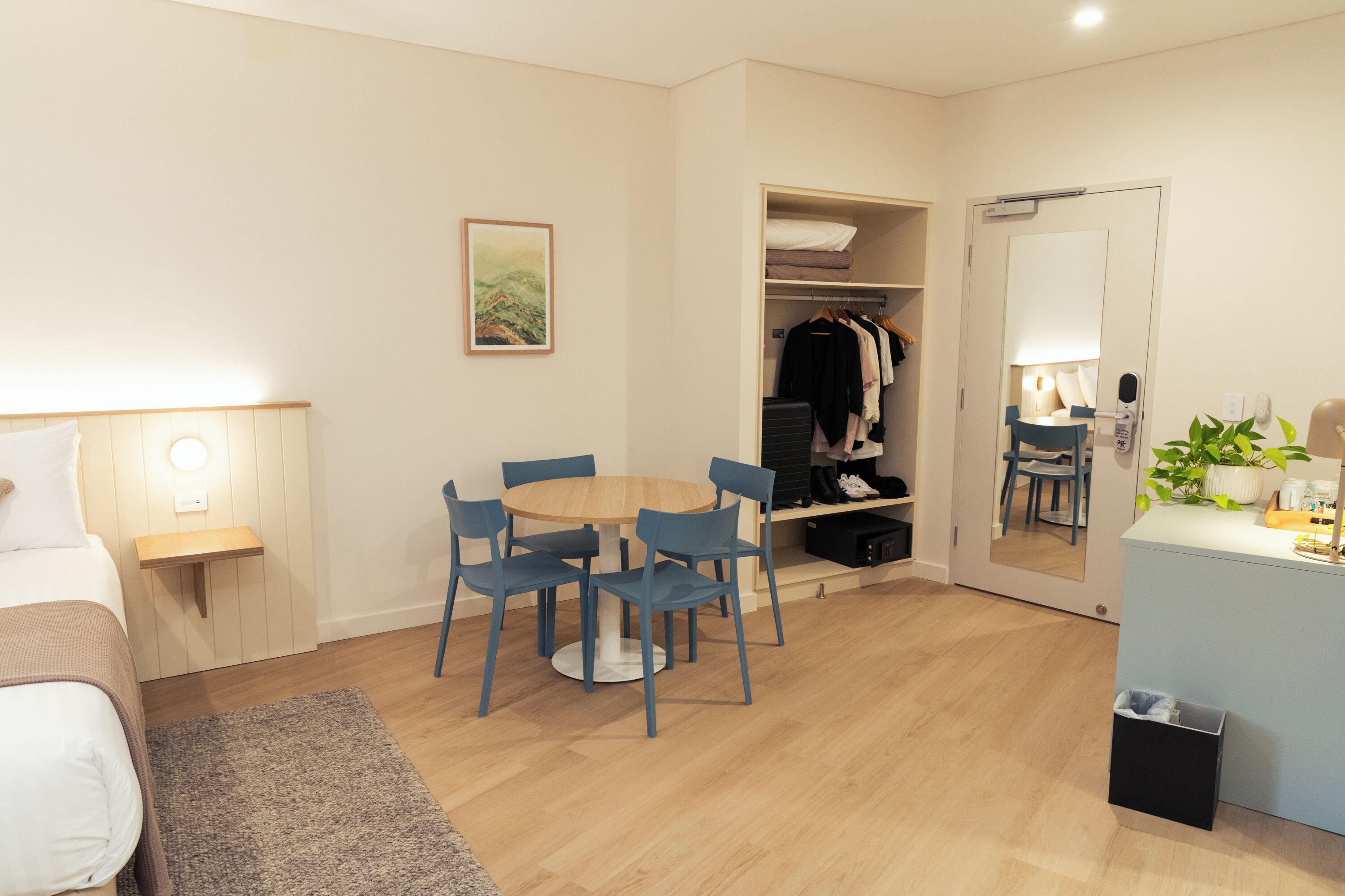 Quarto de hotel com uma cama, uma mesa redonda com cadeiras, um guarda-roupa com roupas penduradas, um espelho e uma pequena planta sobre um balcão.