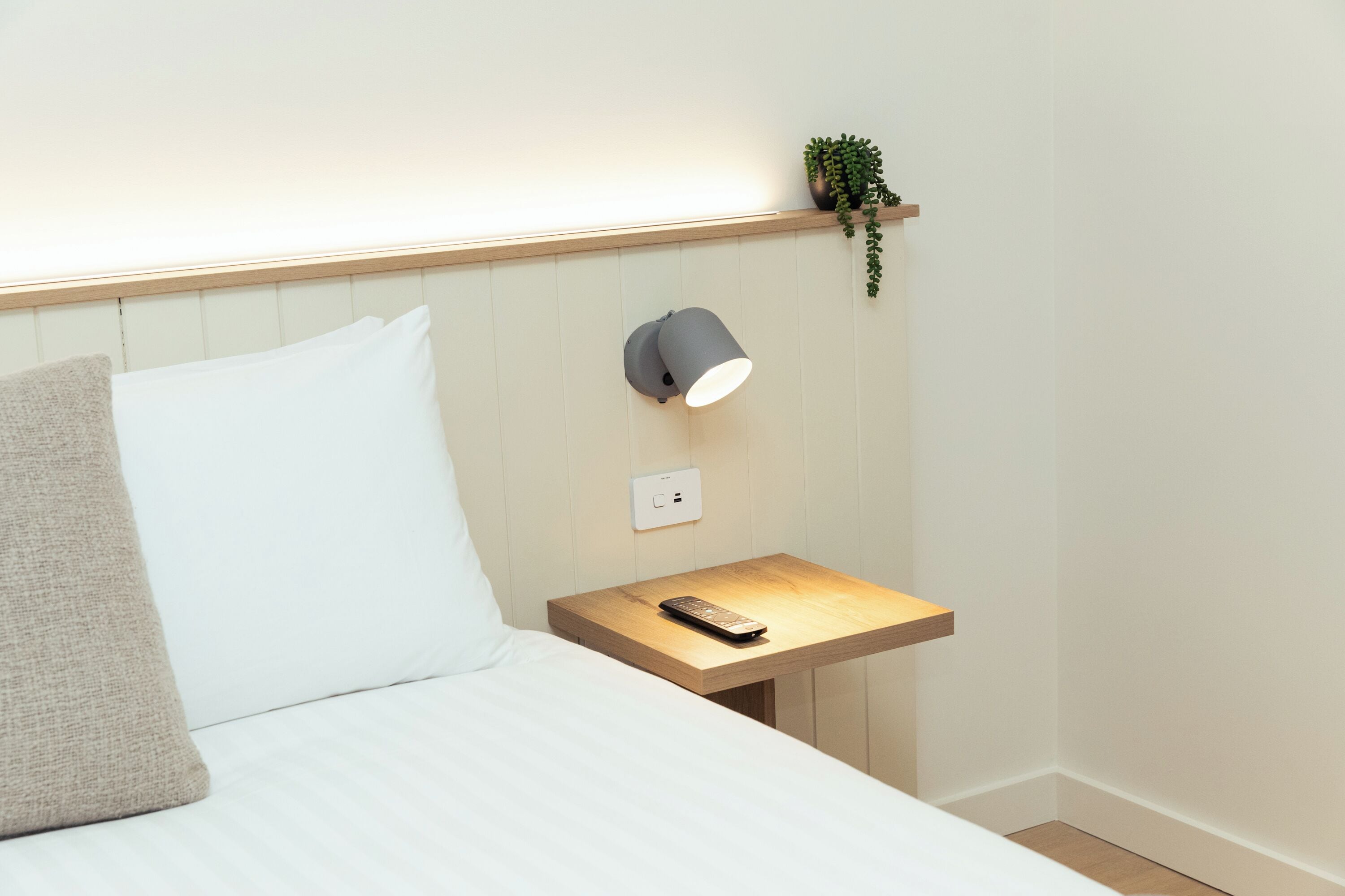 Una cama bien hecha con una almohada, una mesita de noche con un control remoto, una lámpara de pared y una pequeña planta en un estante.
