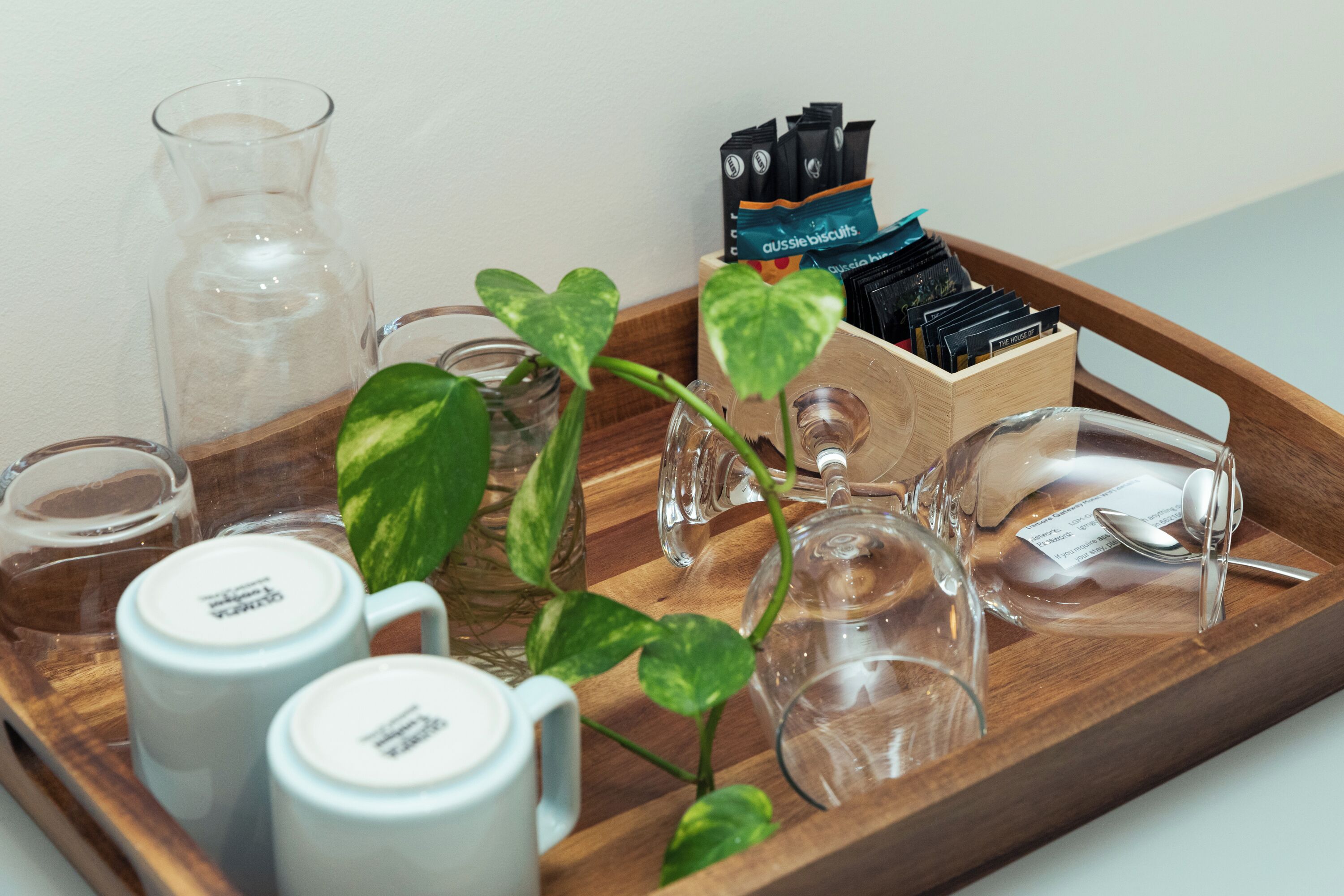 A tray with a glass pitcher, two white mugs, a glass carafe, a small glass, a wooden box with black and blue items, and a potted plant.