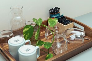 Una bandeja con una jarra de vidrio, dos tazas blancas, una jarra de cristal, un vaso pequeño, una caja de madera con objetos negros y azules, y una planta en maceta.