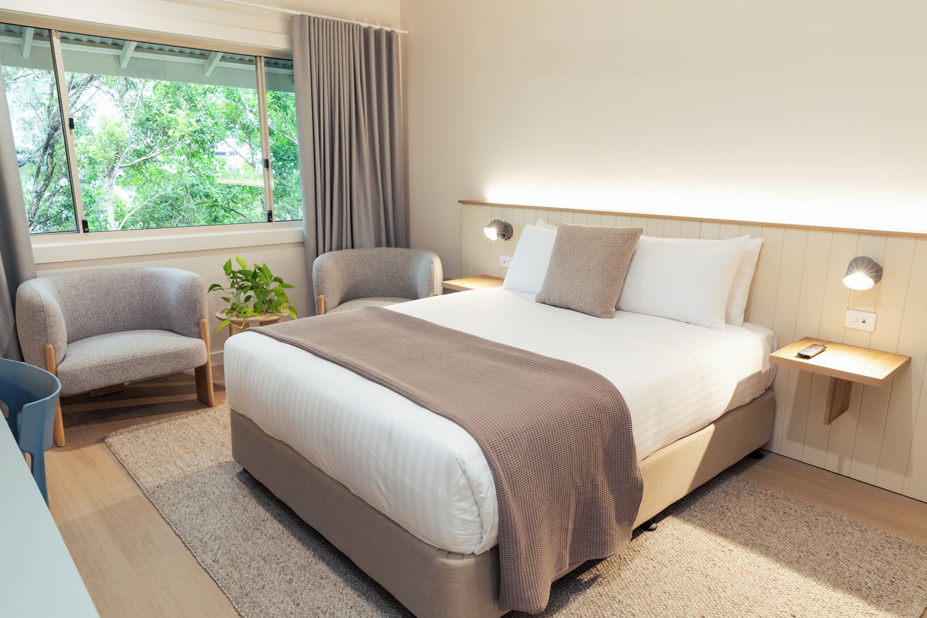 A modern hotel room with a large bed, two armchairs, a small table, and a window with curtains.