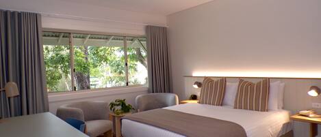 A hotel room with a bed, two armchairs, a small table, a ceiling fan, and a window with curtains.