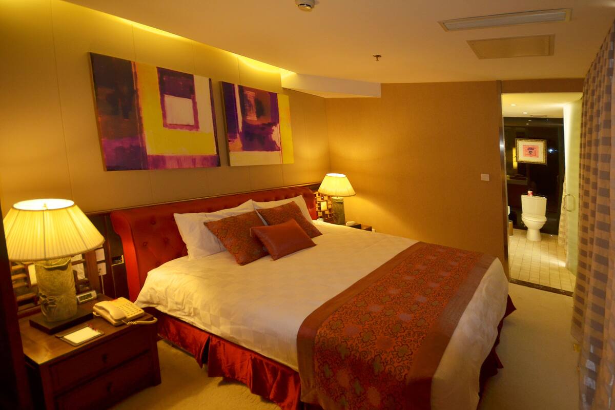 Una habitación de hotel con una cama grande, dos lámparas de noche, una mesita de noche con un teléfono, y un baño visible a través de una puerta abierta.