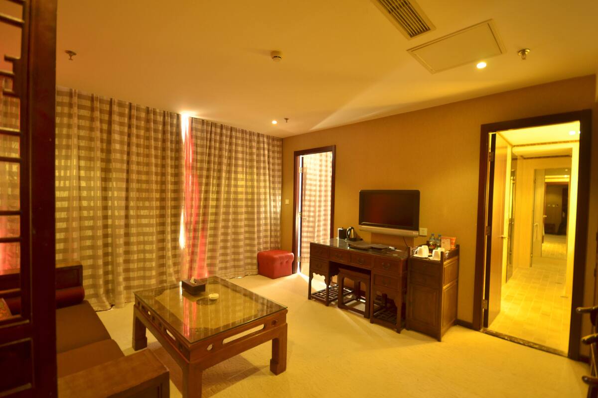 Habitación de hotel con una mesa de centro de vidrio, un televisor sobre un mueble de madera, un escritorio con una silla y un reposapiés rojo.