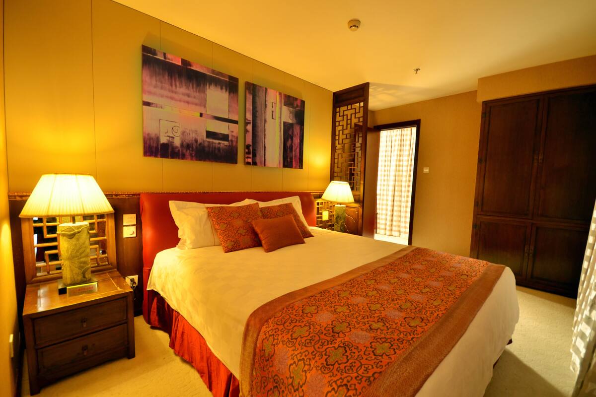 Una habitación de hotel con una cama grande, dos mesitas de noche con lámparas, un cabecero de madera y una obra de arte de gran tamaño sobre la cama.